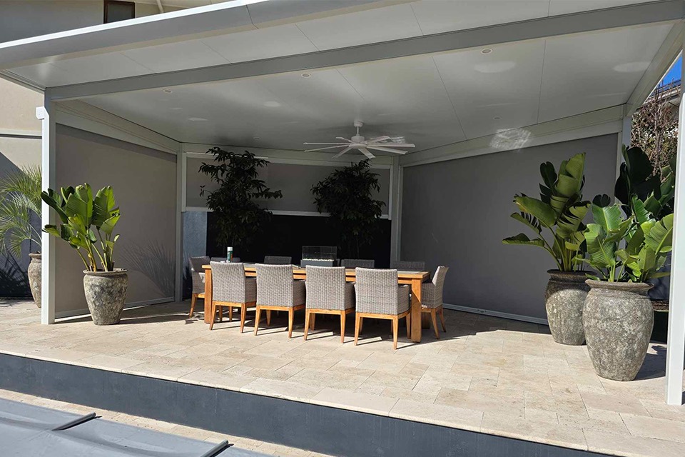 Insulated patio with outdoor blinds and a ceiling fan over a paved dining area with pot plants