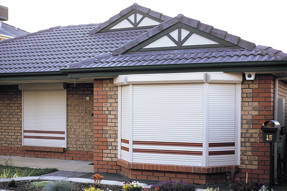 Cream roller shutters lowered over a bay window and a front window of a brick home