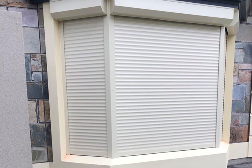 Cream roller shutters fitted to each pane of a bay window in a stone wall