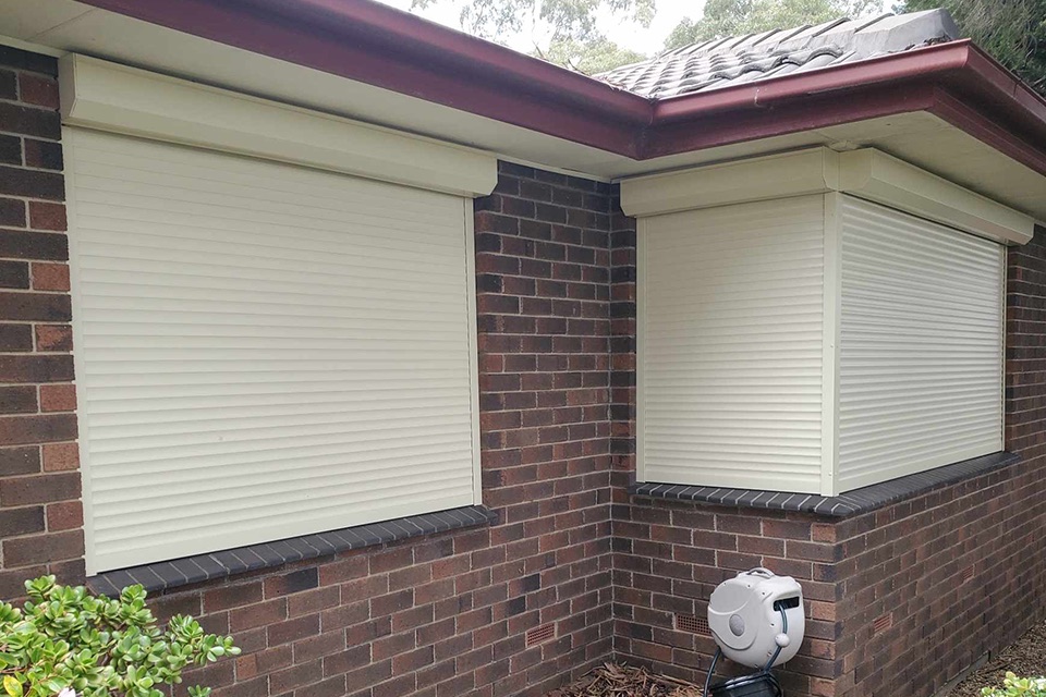 Cream roller shutters on two windows at the corner of a brick home