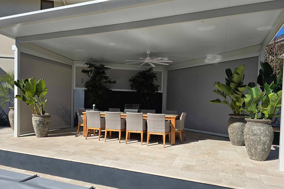 Grey outdoor blinds on an insulated patio with a ceiling fan over a long outdoor dining table