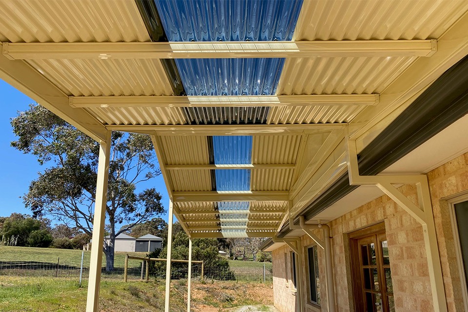 Cream steel pergola roof with polycarbonate strips letting light through, beside a brick home